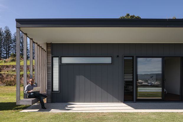 Exterior view of a compact contemporary single-storey home with a flat roof, dark vertical cladding, broad roof overhang, and floor-to-ceiling glazing. A slim covered outdoor seating nook is integrated along the facade, with a person seated on a built-in bench. The property sits in a landscaped suburban or semi-rural setting, and the glazing reflects a distant water or valley view.