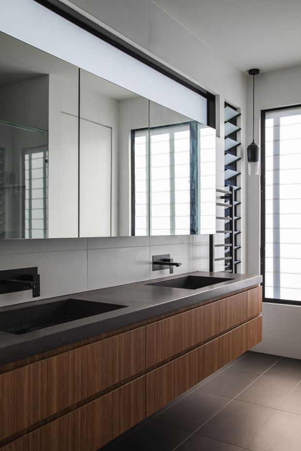 A contemporary bathroom featuring a long floating double vanity with dark stone benchtop, integrated rectangular basins, wall-mounted black tapware, and warm timber-grain cabinetry. Large mirrored wall cabinets reflect bright windows, while black-framed windows, a vertical open shelf, pendant light, and large grey floor tiles complete the clean, high-contrast design.