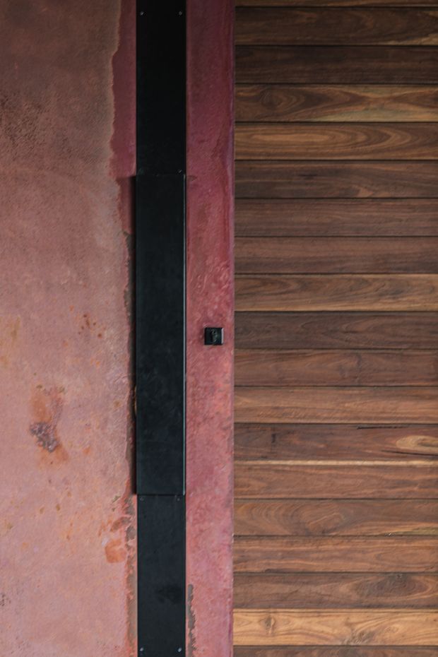 Close-up architectural detail of a dark horizontal timber-clad door or wall panel beside a rough, mottled reddish earthen or plaster wall. A tall narrow black metal vertical strip with visible fasteners and a small black switch or latch forms the junction between the two surfaces. The image emphasises contrasting timber, metal, and textured mineral finishes.