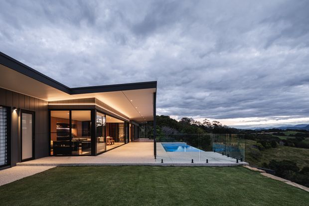 A contemporary single-storey house viewed from the exterior at dusk, featuring a sharply angled flat roof, dark vertical cladding, extensive floor-to-ceiling glazing, and a broad covered terrace. A small swimming pool with glass balustrades sits beside the home, overlooking a broad rural or semi-rural landscape. Warm interior lighting contrasts with the cool overcast sky.