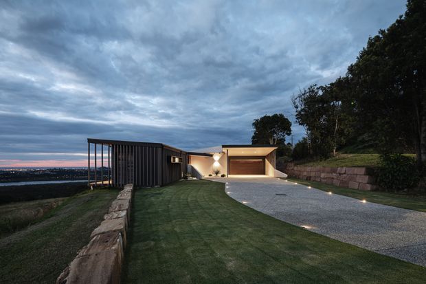 Twilight exterior view of a contemporary single-storey residence on a sloped site. The home features a long dark vertical-clad pavilion with an open framed end, a pale rendered central entry volume, and a large integrated garage with a warm timber-toned door. A broad gravel driveway curves through the landscaped lawn, with low retaining walls and discreet ground lighting. Distant urban or coastal lights are visible beyond the site.