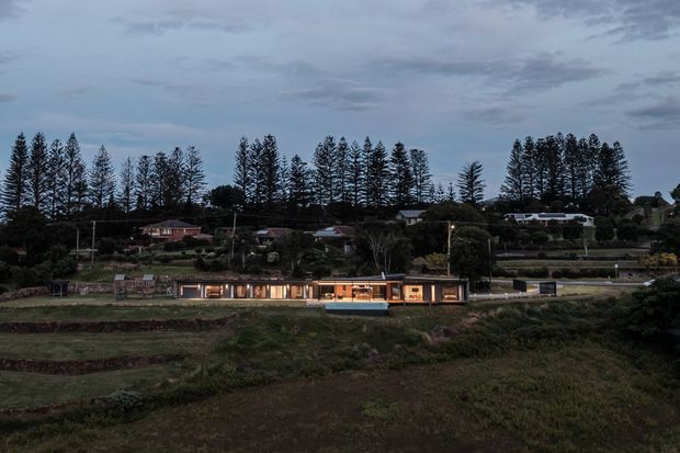 Twilight exterior view of a long, low modern residence set into a landscaped, semi-rural hillside. The house has extensive warm-lit glazing, a dark linear facade, and a small rectangular pool or water feature in front. Tall narrow evergreen trees and neighboring homes form the backdrop.