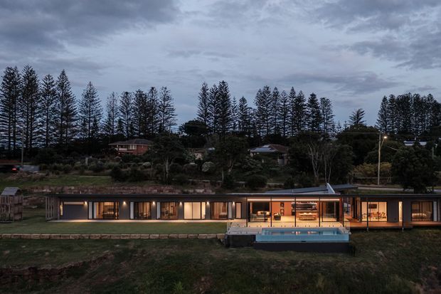 Twilight exterior view of a long, low-slung single-storey residence set into a landscaped, tree-lined site. The house has extensive floor-to-ceiling glazing, dark slender window frames, a shallow flat or low-pitched roof, and warm interior lighting visible through the windows. A raised rectangular swimming pool and terrace sit directly in front of the central living area.