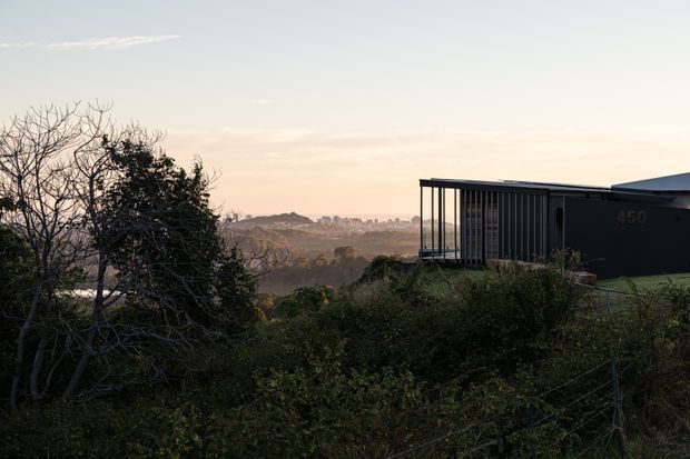 Exterior view of a dark, contemporary flat-roofed building set on a vegetated elevated site, with extensive vertical screening and a covered glazed terrace or balcony. A broad distant urban landscape is visible beyond the surrounding trees and shrubs in soft evening light.