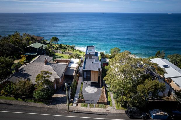 Elevated aerial view of a coastal residential neighbourhood, showing several modern homes on a steep site overlooking a bright blue ocean. The central property has a narrow contemporary form, flat or low-pitched roof sections, a dark facade, landscaped entry path, and a rooftop terrace or upper-level outdoor area. Dense vegetation and neighbouring houses frame the ocean outlook.