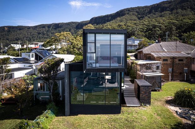 Exterior view of a contemporary multi-level residential home in a suburban hillside setting. The house features a dark-framed glazed upper volume, extensive floor-to-ceiling glazing, dark cladding, and a flat roof, with neighbouring homes and a forested ridge behind it.