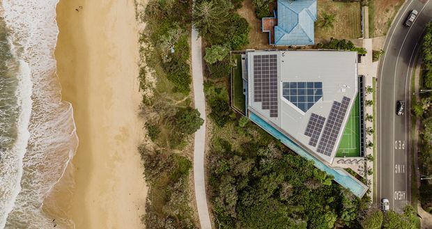 Aerial view of a modern coastal property directly beside a sandy beach. The large angular-roofed building has extensive rooftop solar panels and a small rooftop green recreation area, surrounded by dense coastal landscaping, with a road running along the opposite side.