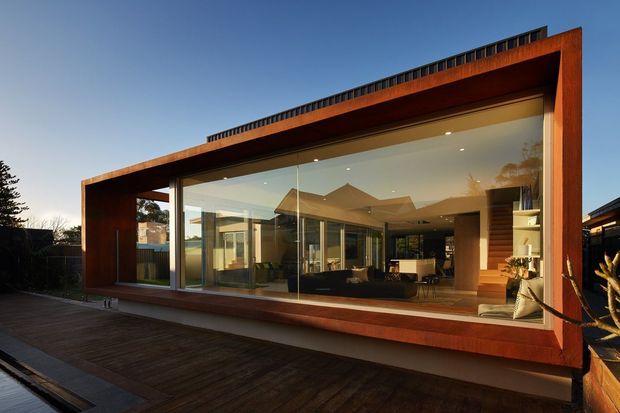 Exterior view of a contemporary single-storey pavilion-like house, framed by extensive floor-to-ceiling glazing and warm rust-toned cladding. The building opens onto a broad timber deck, with a visible open-plan living and dining area inside. A dark roof edge and raised rooftop element emphasize the strong horizontal form.