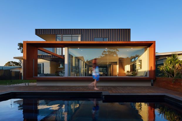 Front exterior view of a modern multi-level residence beside a swimming pool. The composition is dominated by a large glazed pavilion framed in warm-toned timber or rust-coloured metal, with an upper box clad in dark vertical ribbed panels. Floor-to-ceiling glazing reveals a warm contemporary interior, while the pool and timber deck occupy the foreground.