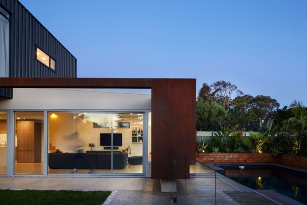 Twilight exterior view of a contemporary multi-level residence with dark vertical cladding, a large timber-framed glazed living area, and a warm-toned timber-clad portal structure. The open-plan living room is visible through the glazing, with a dark sofa, wall-mounted television and built-in shelving. A narrow reflecting or swimming pool sits beside a paved terrace with glass balustrading and integrated raised garden beds.