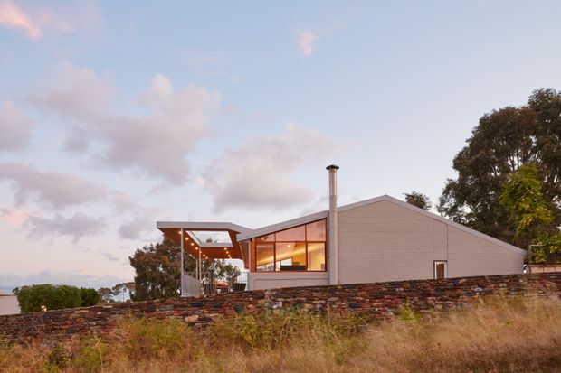A low, single-storey contemporary house viewed from outside at dusk. The home has a prominent pitched gable roof, pale horizontal cladding, extensive warm-lit glazing, a projecting covered outdoor terrace or entry structure, and a tall metal chimney. A layered stone retaining wall runs across the foreground, with mature trees and dry grasses surrounding the property.