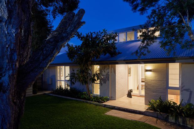 Twilight exterior view of a low-rise contemporary house, with a broad corrugated metal roof, light rendered or masonry walls, extensive warm-lit glazing, and a raised clerestory-like upper section. A landscaped lawn, mature tree, paved path, and planted garden beds frame the facade.