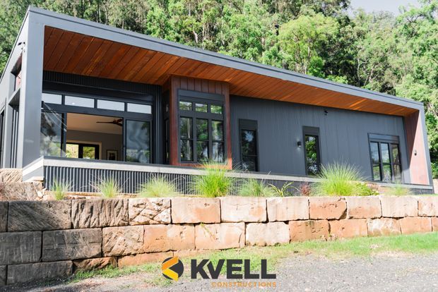 Single-storey contemporary house set into a wooded, sloping site, with a dramatic low-pitched roof and deep timber-lined soffit. The dark grey facade combines vertical cladding with large black-framed windows and doors, opening toward a raised terrace edged by a metal balustrade. A substantial rough-stone retaining wall defines the foreground.