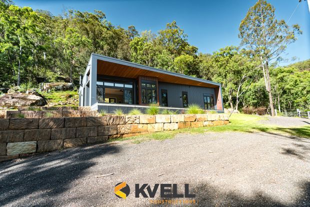 A compact single-storey contemporary dwelling set among dense Australian bushland. The building has a long, low form with dark grey vertical cladding, extensive glazing, a sloping roof with a warm timber-lined soffit, and substantial layered sandstone retaining walls in front. A gravel driveway and landscaped grass surround the home.