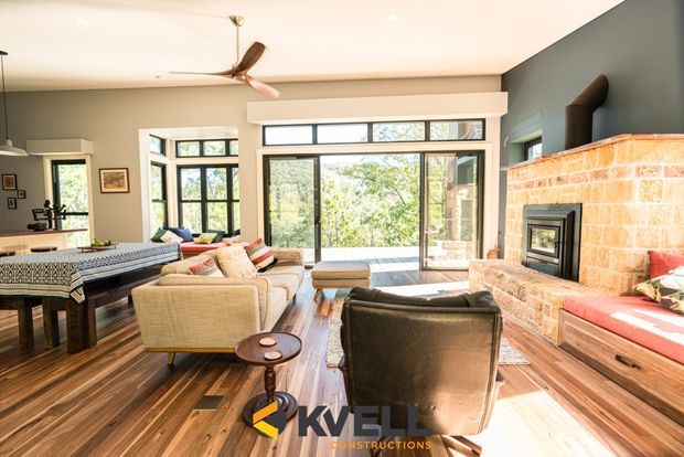 Bright, open-plan living room with warm timber flooring, a large black-framed glazed opening to the outdoors, neutral upholstered seating, a dining table, and a prominent exposed brick fireplace with integrated wood-burning stove. The room has a contemporary yet warm character and abundant natural light.
