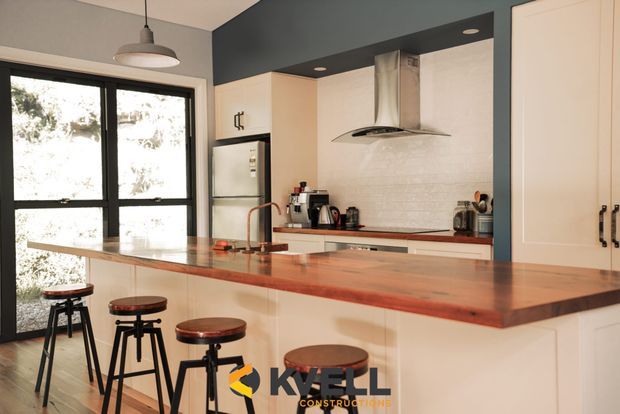 A bright contemporary kitchen with a large peninsula island and dark timber benchtop, fitted with cream cabinetry, black-framed glazed doors, a stainless-steel rangehood, and a pale tiled splashback. Timber-topped stools line the island, while a pendant light and dark ceiling bulkhead add contrast.