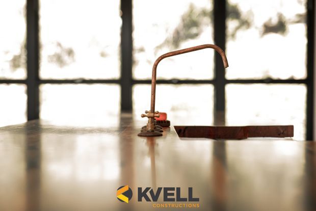 Close-up of a distinctive copper-toned gooseneck tap or pipe fixture mounted on a polished countertop, with a long dark wooden or metal lever beside it. Large black-framed industrial-style windows fill the background, creating a warm, workshop-like architectural setting. Kvell Constructions branding is visible at the bottom.