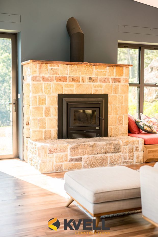 Bright interior view focused on a substantial masonry fireplace with a black metal wood-burning insert and flue. The fireplace is constructed from warm, roughly textured light sandstone or limestone blocks, with an integrated hearth bench. Timber flooring, large windows and doors, and a built-in window seat with red cushions are visible, along with a neutral upholstered ottoman in the foreground.