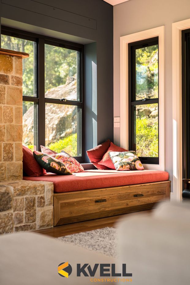 Warm interior detail featuring a built-in window seat with red upholstered cushioning and colourful patterned pillows, integrated timber drawers, dark-framed windows, and a stone-clad fireplace or masonry element at left. Sunlight filters through the windows, with a natural rocky landscape visible outside. A construction company watermark appears at the bottom.