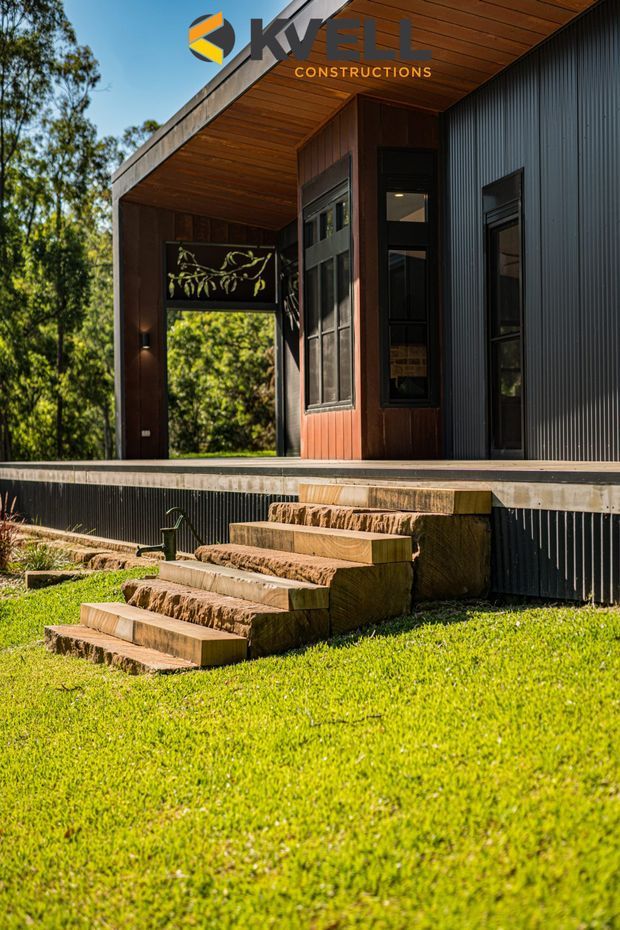 Exterior view of a contemporary dark-clad house set in a leafy landscape, showing a covered entry or terrace with timber soffit, black corrugated wall cladding, tall windows, and a stepped rough-sawn timber and stone stairway leading up from the lawn. A construction company logo is overlaid near the top of the image.