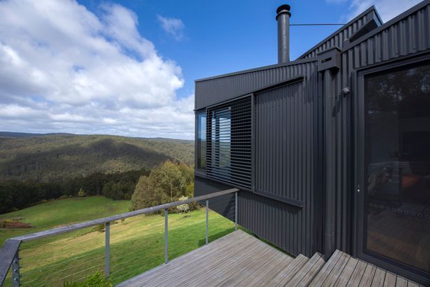 Exterior view of a compact modern dwelling on a raised timber deck, overlooking rolling forested hills and open rural landscape. The building has dark corrugated metal wall cladding, a steeply angled roof form, black-framed glazing, external louvres, a chimney, and a glass balustrade around the deck. The image is primarily architectural and showcases the elevated outdoor terrace and rugged contemporary form.