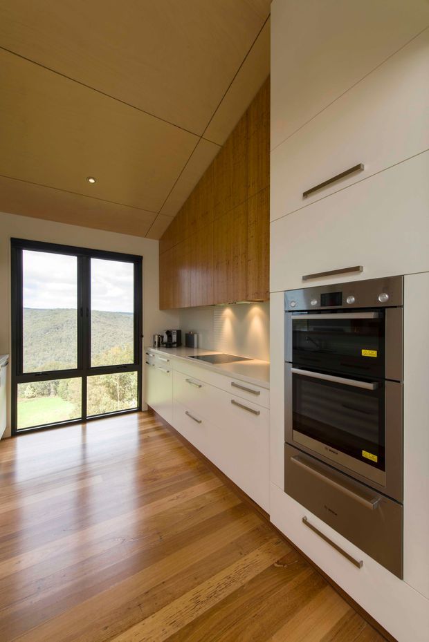 A compact, contemporary kitchen with white handleless-style cabinetry, integrated stainless-steel double ovens, a pale stone benchtop, and a warm timber overhead cabinet wall. The room has polished timber flooring, a timber-lined ceiling with recessed lighting, and large black-framed glazing overlooking a green landscape.