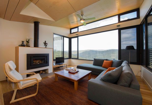 A warm, contemporary living room with a large grey sectional sofa arranged around a timber coffee table and a built-in fireplace. Full-height corner glazing and clerestory windows provide expansive views, while a timber-panelled ceiling, dark window frames, and a ceiling fan add architectural character. A white lounge chair, television, rug, and orange accent cushion complete the inviting interior.