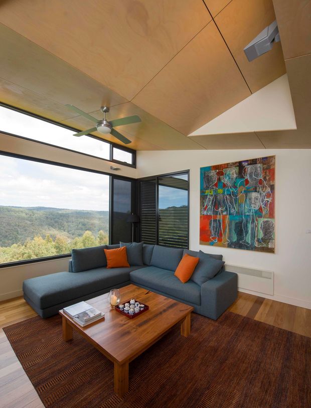 A contemporary living room with expansive corner glazing overlooking a wooded landscape. The room features a large dark blue-grey sectional sofa with orange cushions, a solid timber coffee table, a large textured brown rug, colourful abstract wall art, timber flooring, and a warm timber-lined ceiling with a ceiling fan. A high-level window band and openable louvred glazing add architectural interest and natural light.