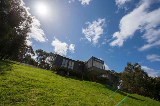 Low-angle exterior view of a dark, modern elevated house set on a steep grassy hillside, with extensive glazing, angular projecting volumes, slender supporting structure, and surrounding trees beneath a bright blue sky.