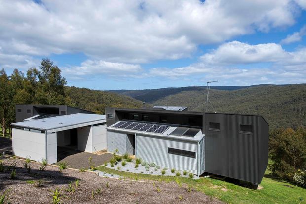 A contemporary two-part residence set on a sloping, landscaped site, with low-profile flat and mono-pitched roof forms, dark ribbed cladding, pale metal wall panels, broad roof-mounted solar panels, and a covered connecting structure. Rolling forested hills are visible behind the building under a partly cloudy sky.