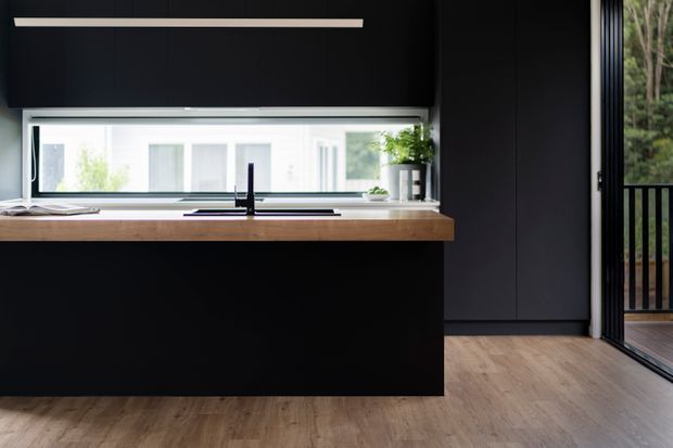 A minimalist modern kitchen with a matte black island and full-height dark cabinetry, featuring a thick timber benchtop, integrated black sink and tap, linear pendant lighting, and a long horizontal window. Timber flooring and an adjoining glazed door open toward a leafy exterior balcony or deck.