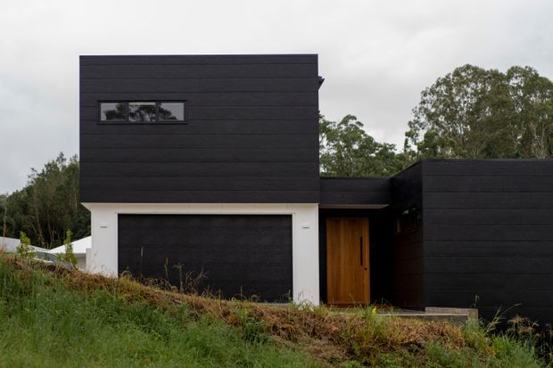 Exterior view of a modern, angular single-storey-to-two-level residential building on a sloping, vegetated site. The facade is clad in dark horizontal boards, with a contrasting white garage surround, narrow horizontal window, and warm timber entry door. The composition has strong rectilinear volumes and a flat roofline beneath an overcast sky.