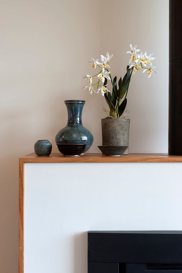 Close-up interior detail of a minimalist fireplace or built-in cabinet feature. A warm timber-edged white surround supports a dark stone or ceramic vase arrangement and potted white flowers, with a black firebox or inset feature visible below. The composition is softly lit and restrained, with a neutral wall backdrop and a refined, calm aesthetic.