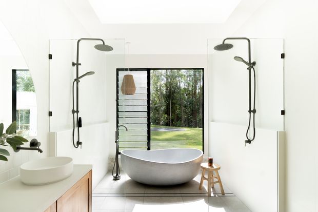 Bright contemporary bathroom with twin open showers enclosed by clear glass screens, a freestanding oval bathtub, vessel basin on timber joinery, and large windows opening to a leafy outdoor setting. The room has white surfaces, dark shower fittings, pale flooring, and abundant natural light.