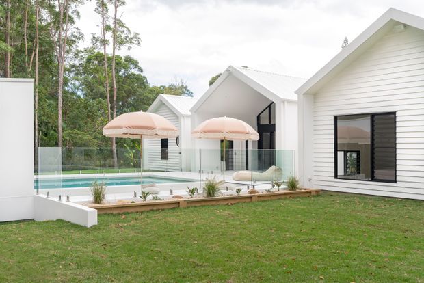 Exterior view of a contemporary white-clad residential property composed of interconnected pitched-gable volumes. The landscaped lawn leads to a glass-fenced swimming pool and a paved outdoor entertaining area with pale blush umbrellas. Black-framed glazing, white horizontal weatherboard cladding, and a light metal roof create a crisp, resort-like presentation, set against mature trees.
