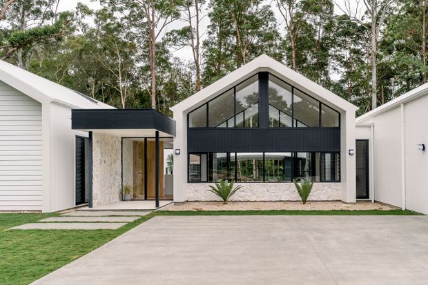 Front exterior view of a contemporary single-storey residence with a steep gable roof, dark-framed floor-to-ceiling glazing, white rendered or clad walls, and a pale stone feature wall beneath the windows. The home is set among mature trees, with a broad concrete driveway, lawn, and integrated side structures visible.