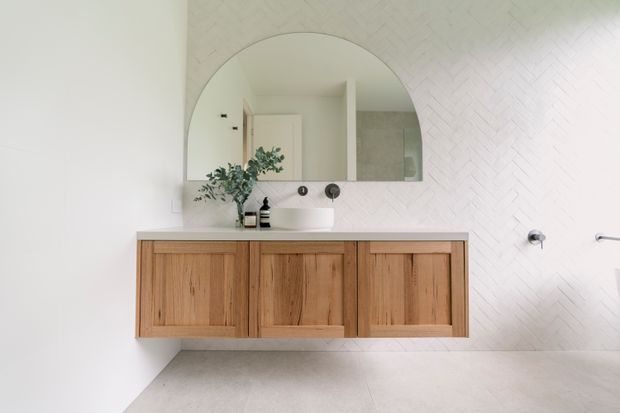 Bright contemporary bathroom vanity with a wall-mounted natural timber cabinet, white vessel basin, large arched mirror, and pale herringbone wall tiling. Minimal black tapware and small greenery add restrained contrast.