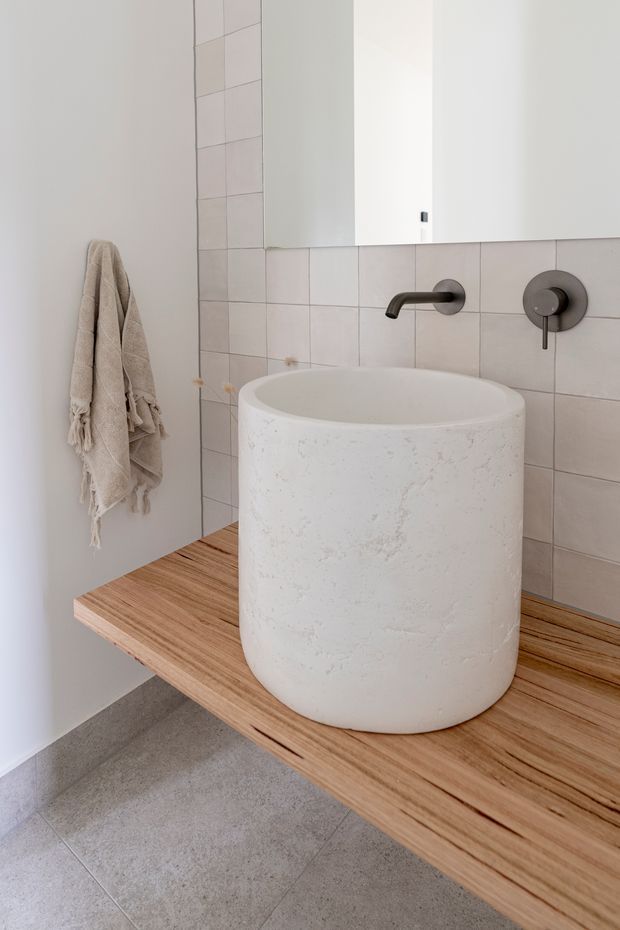 A minimalist bathroom vanity featuring a large round white vessel basin on a floating timber countertop. Pale square wall tiles, a large mirror, matte dark wall-mounted tapware, and a neutral textured towel create a restrained, warm contemporary interior.