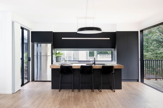 Bright contemporary kitchen with a black island and full-height cabinetry, stainless-steel refrigerator, pale timber flooring, white benchtops, and three black bar stools. Large windows and glazed doors provide views to a leafy exterior balcony or deck, while a black pendant light hangs above the island.