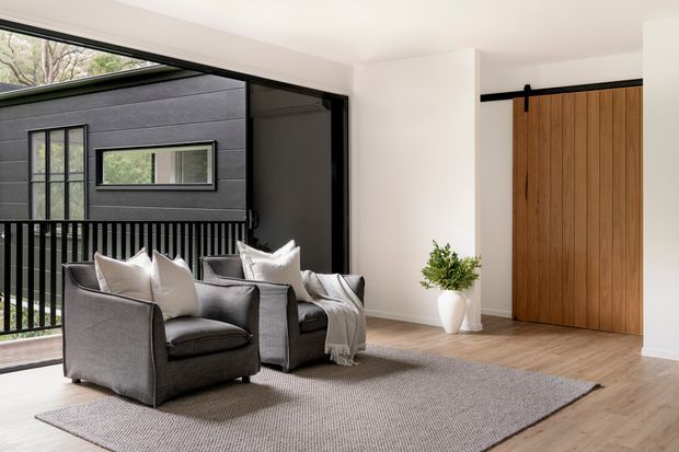 A bright, contemporary living room or entry-level lounge with two grey upholstered armchairs, a large neutral rug, and a small white side table with greenery. Full-height black-framed glazing opens to a balcony or exterior walkway, while a large vertical timber sliding door with black hardware forms a strong feature at the rear. Light timber flooring, white walls, and a soft neutral palette create a calm, refined atmosphere.