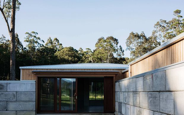 A low, modern building set within a wooded landscape, viewed from an enclosed courtyard. The foreground is defined by grey concrete block walls, with a timber-clad upper wall and a shallow corrugated metal roof. Dark timber-framed glazed doors open into a shadowed interior, while trees and distant hills are visible beyond.
