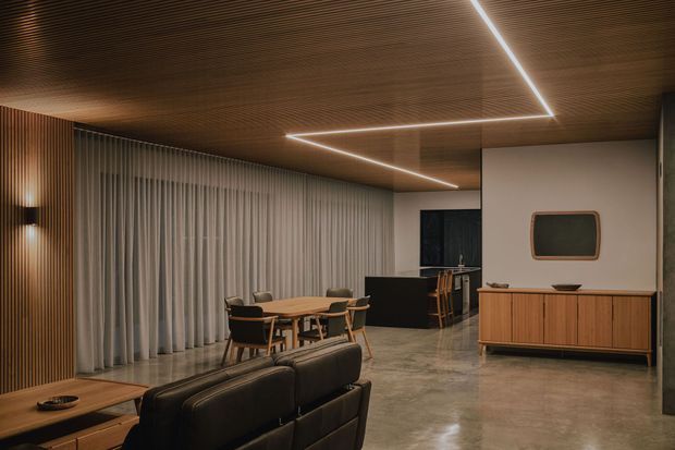 A sophisticated open-plan living and dining area with a low black leather sofa in the foreground, a timber dining table and upholstered chairs, and a timber sideboard against a pale wall. Full-height sheer curtains screen the glazing, while a dark kitchen or bar zone sits beyond the dining area. The space features a richly slatted timber ceiling with integrated linear LED lighting, vertical timber detailing, and a polished grey concrete floor, creating a warm, refined contemporary interior.