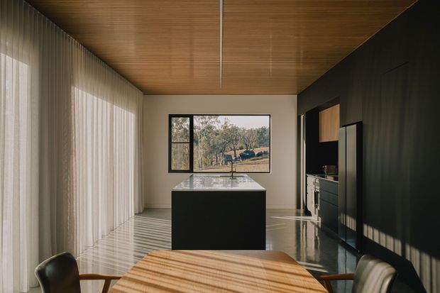 A refined contemporary kitchen and dining area with a large dark island, integrated cabinetry along a black feature wall, and a timber dining table in the foreground. Full-height sheer curtains line one side, while a warm timber-lined ceiling adds natural texture. The space is illuminated by strong filtered daylight, with a framed landscape visible through the rear window.