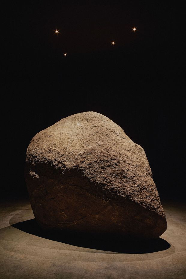 A large irregular natural boulder is dramatically spotlighted in a very dark, gallery-like interior. The rock sits on a smooth floor and casts a strong shadow, while several small star-like ceiling lights glow above. The image is minimalist, moody, and sculpture-focused.