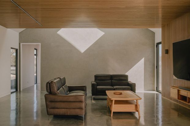 A refined modern living room with a pair of dark leather sofas arranged around a low timber coffee table. The room features a polished concrete floor, warm timber-lined ceiling and TV wall, smooth neutral plaster walls, a slim linear ceiling light, and strong geometric sunlight patterns. Floor-to-ceiling glazing and a glazed door bring in natural light, creating a calm, minimalist atmosphere.
