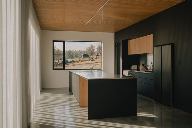 A refined contemporary kitchen with a large central island, dark charcoal cabinetry, warm timber overhead cabinetry and ceiling lining, and a pale stone benchtop. Full-height sheer curtains and expansive black-framed glazing bring filtered daylight into the space, with a dry rural landscape visible outside. The room has a clean, minimalist composition and strong contrast between the dark joinery and warm timber surfaces.
