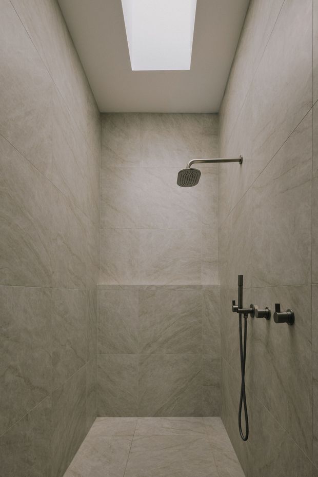 A compact contemporary walk-in shower enclosed by floor-to-ceiling pale stone-look tiles. The shower has dark brushed-metal fixtures, including a square rainfall showerhead, handheld hose, and wall-mounted controls. A large rectangular skylight or ceiling light brightens the otherwise minimal, warm-grey space.