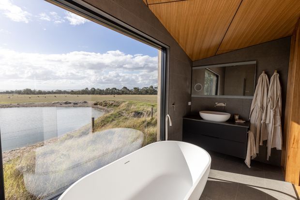 A contemporary bathroom with a freestanding white bathtub positioned beside expansive floor-to-ceiling glazing overlooking a rural waterway and open landscape. The room features dark grey tiled walls and floor, a timber-lined ceiling, dark built-in vanity cabinetry with a white vessel basin, frameless mirror, and warm timber detailing. Neutral robes hang beside the vanity.