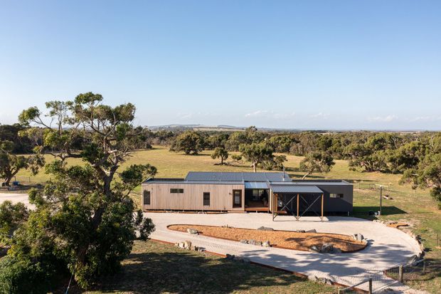 A single-storey contemporary rural home set in an expansive open landscape. The building has a low-pitched metal roof, dark and timber-toned exterior walls, broad glazing, and a covered entry or outdoor living structure. A pale curved driveway and landscaped gravel beds surround the house, with scattered mature trees and farmland extending to the horizon.