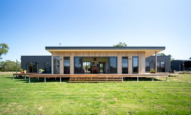 Single-storey contemporary house viewed from the front across a lawn. The home has a long low-profile form with a flat roof, dark exterior wall sections, warm vertical timber-look cladding around the central glazed living area, extensive floor-to-ceiling windows and doors, and a raised timber deck with steps and slim supports.